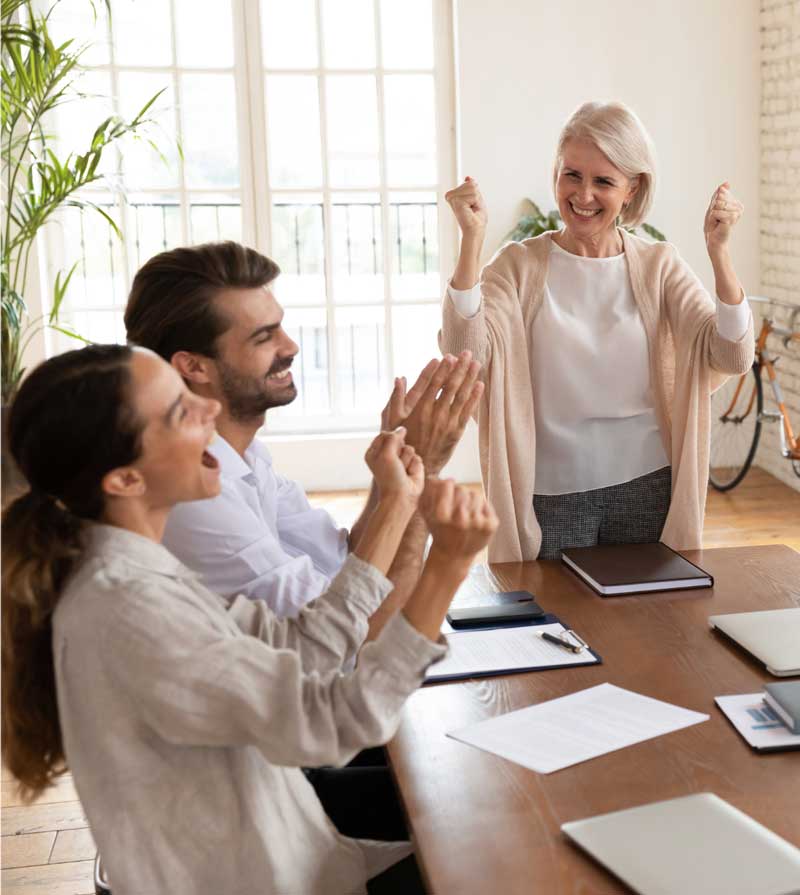 A group of three people at a table celebrate enthusiastically, showing excitement during a meeting, with a bright, cheerful atmosphere and natural lighting.