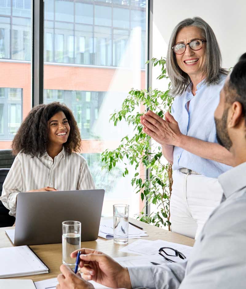 A woman stands in a bright office, engaging with two seated colleagues. They appear to be in a meeting, discussing ideas, with plants and large windows in the background.