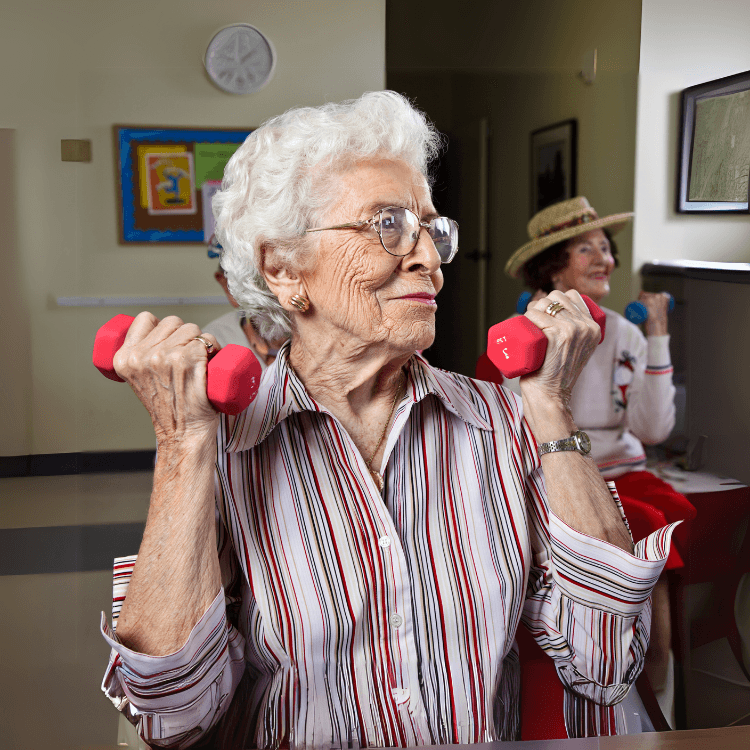 an elderly woman with short hair and a striped dress shirt sits at a table with elbows bent up while holding a small hand weight in each hand, there is another woman exercising while sitting at a table in the background, and they appear to be a a multi-purpose room- the point of the photo is to highlight the benefits of movement at all ages